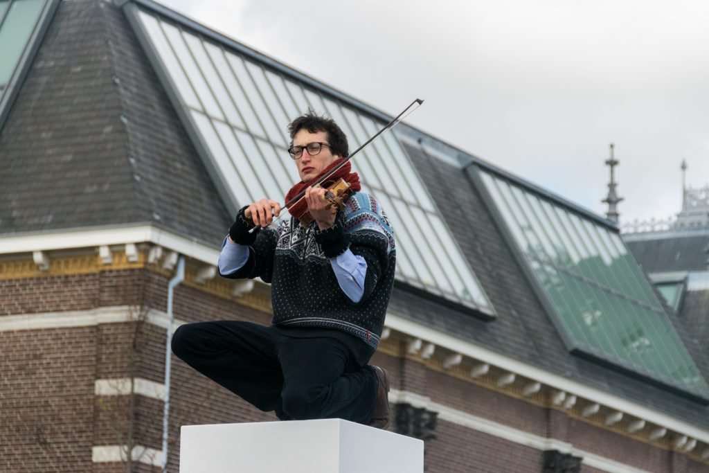 Knielend op een van de I AMsterdam-letters speelt een muzikant zijn viool voor het toeristenpubliek. Foto: Lucas Faijdherbe